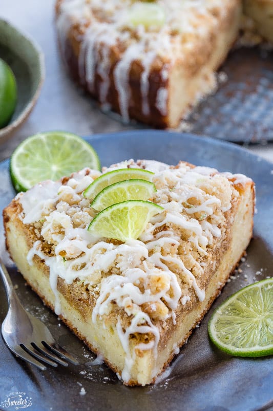 A slice of Key Lime Coffee Cake with streusel topping, white chocolate drizzle and lime garnish on a plate with a fork