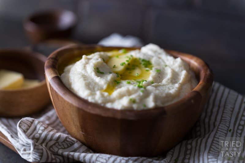 Serving of cauliflower mash in a wooden bowl topped with two pats of butter & chopped chives